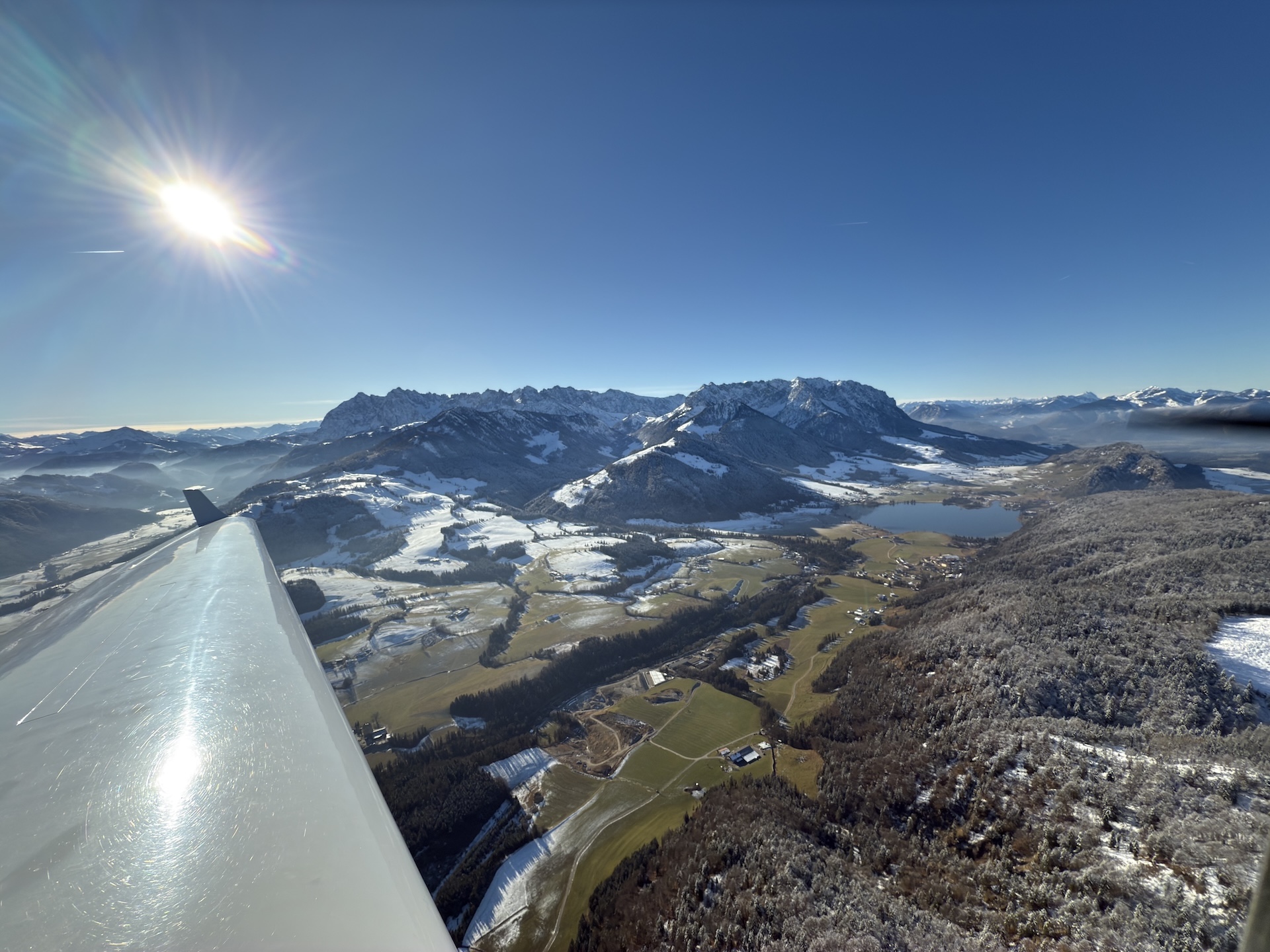 Das Bild zeigt einen Blick aus einem Flugzeug über das winterliche Kaisergebirge mit dem markanten Wilden Kaiser und dem sanfteren Zahmen Kaiser. Im Vordergrund schimmert der Flügel des Flugzeugs, während die tiefstehende Sonne das verschneite Panorama in warmes Licht taucht. Unten erstrecken sich Täler, Wiesen und ein idyllischer See.