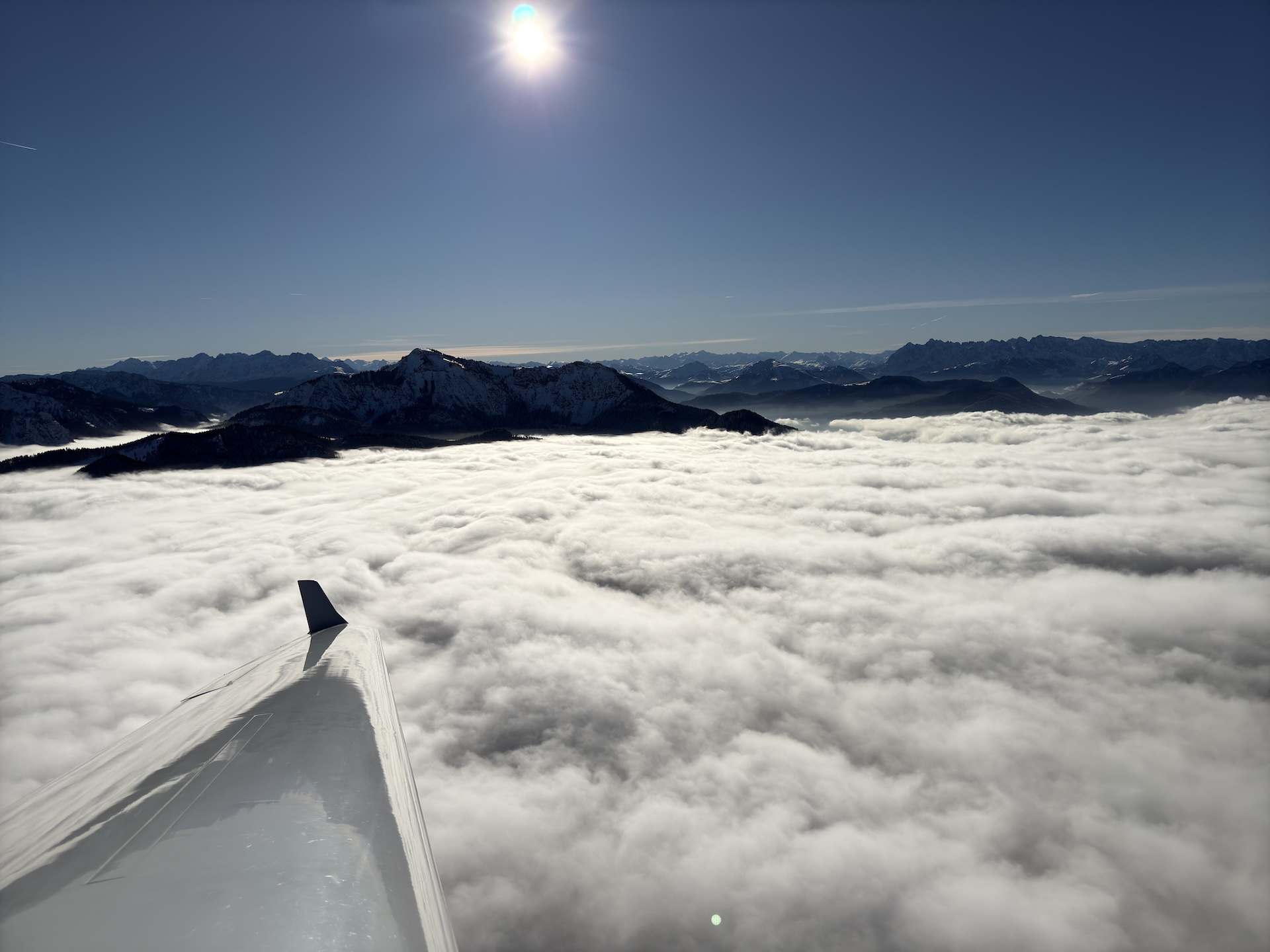 Das Bild zeigt einen atemberaubenden Blick vom südlichen Chiemgau in Richtung Hochalpen. Über einem endlosen Meer aus weißen Wolken ragen dunkle Berggipfel hervor, während die tiefstehende Sonne den klaren Himmel erleuchtet. Im Vordergrund ist die glänzende Tragfläche eines Flugzeugs zu sehen, die die Perspektive eines Flugs über diese majestätische Landschaft unterstreicht.