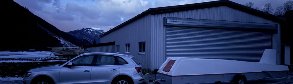 Das Bild zeigt ein Auto, das einen aerodynamischen Anhänger zieht, vor einem Hangar der Segelfluggruppe SGSM. Im Hintergrund erstreckt sich der verschneite Flugplatz Unterwössen, eingerahmt von einer majestätischen Bergkulisse mit Blick auf den Geigelstein und Breitenstein. Die Szene findet in der Dämmerung statt und vermittelt eine winterliche, ruhige Atmosphäre.