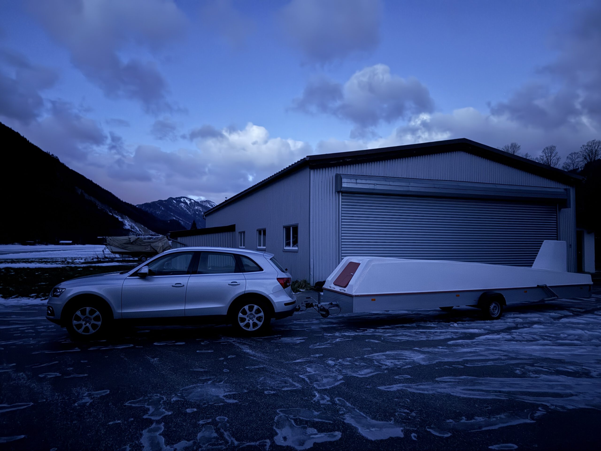 Das Bild zeigt ein Auto, das einen aerodynamischen Anhänger zieht, vor einem Hangar der Segelfluggruppe SGSM. Im Hintergrund erstreckt sich der verschneite Flugplatz Unterwössen, eingerahmt von einer majestätischen Bergkulisse mit Blick auf den Geigelstein und Breitenstein. Die Szene findet in der Dämmerung statt und vermittelt eine winterliche, ruhige Atmosphäre.