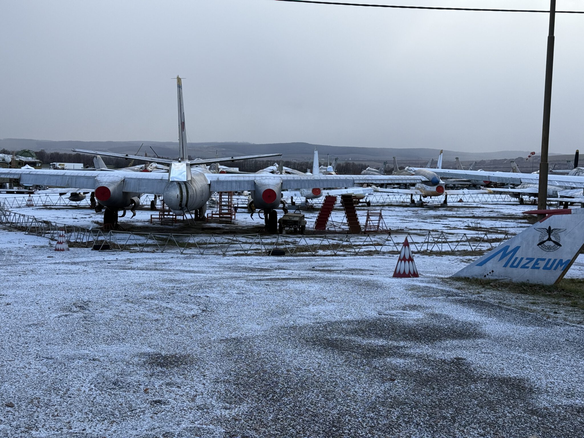 Das Bild zeigt einen verschneiten Außenbereich des Museums in Vyškov, Tschechien. Im Vordergrund steht ein großes, silbernes Militärflugzeug mit markanten roten Abdeckungen an den Triebwerken. Weitere historische Flugzeuge sind in der Hintergrundreihe sichtbar, aufgereiht in einem Freigelände. Eine Tafel mit der Aufschrift “Muzeum” und das offene Gelände mit hügeliger Landschaft ergänzen die winterliche Atmosphäre.