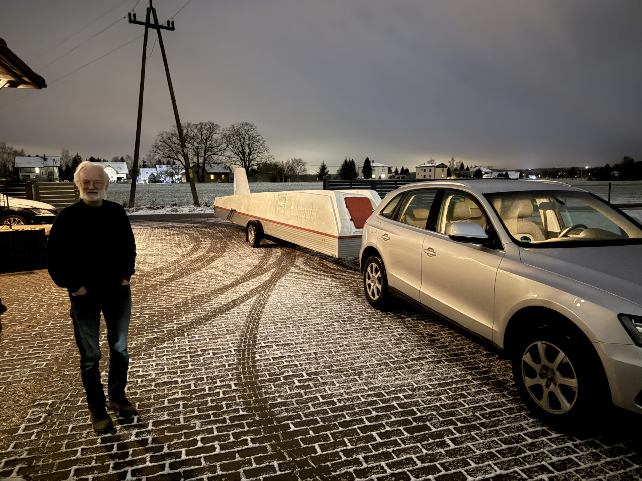 Das Bild zeigt einen Mann mit weißem Haar und Bart, der vor einem silbernen Personenkraftwagen mit einem angehängten Segelfluganhänger steht. Der Boden ist mit leichtem Schnee bedeckt, und Reifenabdrücke zeichnen sich auf den Pflastersteinen ab. Im Hintergrund ist eine winterliche Dorfkulisse mit beleuchteten Häusern.