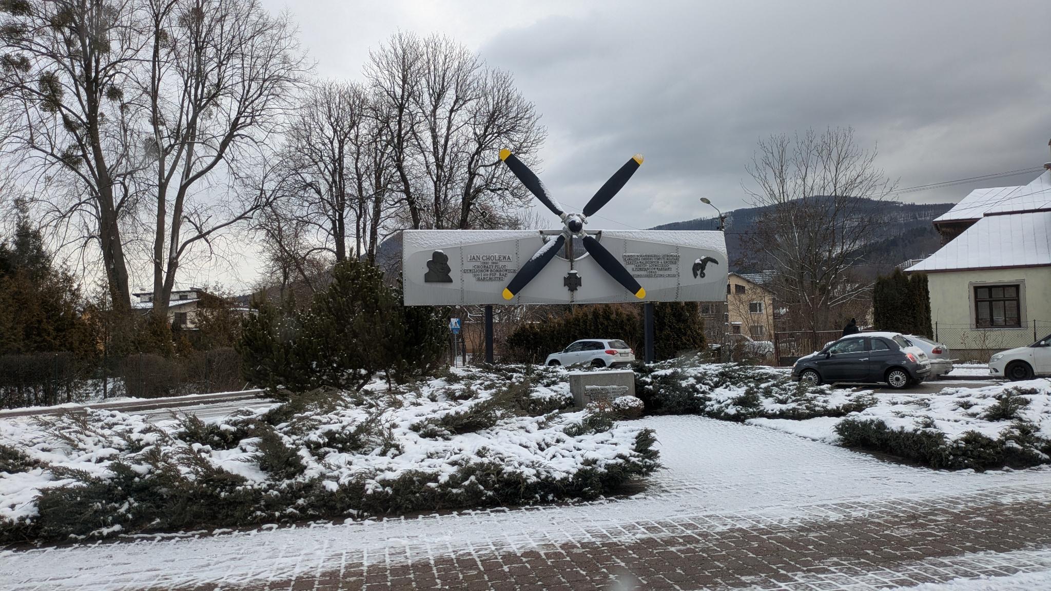 Das Bild zeigt das Fliegerdenkmal in Ustroń, Polen, im Winter. Das Denkmal besteht aus einem Flugzeugpropeller auf einem horizontalen Schild mit Inschriften und Gedenktafeln. Es ehrt Jan Cholewa, einen polnischen Piloten. Umgeben von schneebedeckten Pflanzen und Gebäuden steht es in einer malerischen Landschaft mit kahlen Bäumen und Hügeln im Hintergrund.