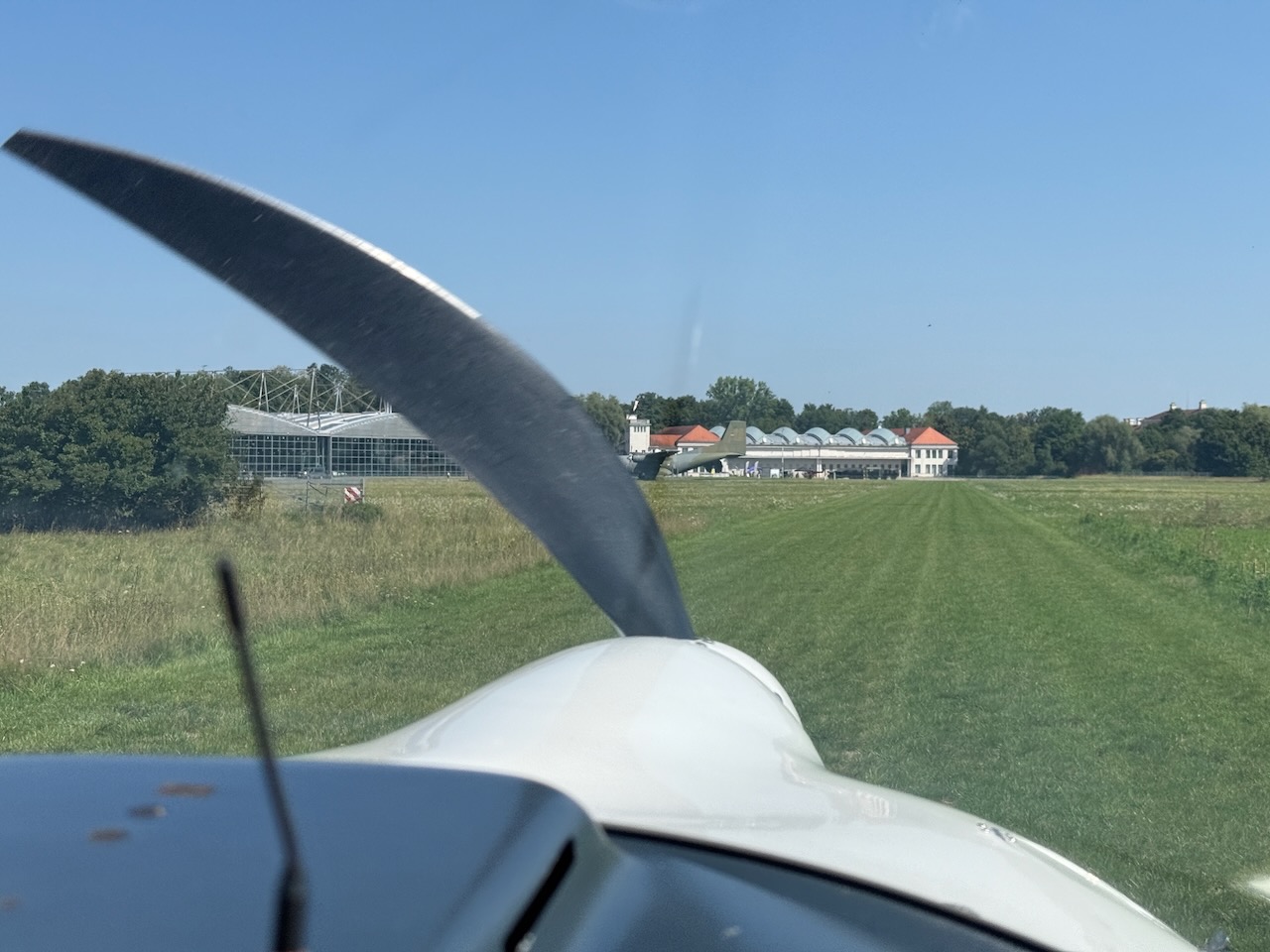 Der Motorsegler Super Dimona HK36 (D-KYSI) am Flugplatz Oberschleißheim Das Bild zeigt einen faszinierenden Moment der Luftfahrtgeschichte aus der Cockpitperspektive: der Blick des Piloten geht nach vorne über den dunkelgrauen Propeller des Motorséglers Super Dimona HK36 mit dem Kennzeichen D-KYSI beim Rollen auf dem Flugplatz Oberschleißheim bei München. Die Aufnahme wurde aus einem begleitenden Luftfahrzeug perspektivisch aufgenommen und präsentiert die charakteristische Propellerform in beeindruckender Detailschärfe. Der Blickwinkel vermittelt authentisch das Gefühl des Piloten während der Bodenrolle. Der Hintergrund offenbart die historische Flugwerft Oberschleißheim des Deutschen Museums mit ihren markanten Hangars und Gebäuden. Die grünen Wiesen und das klare Sommerwetter bilden eine idyllische Kulisse für diese dokumentarische Aufnahme.