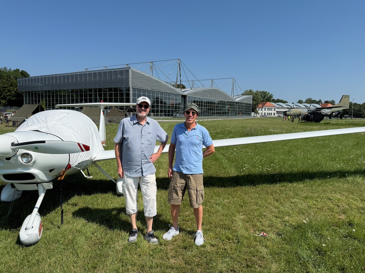 Hans Günther Schreck und Marc Stadtlander der SGSM.de Der Motorsegler Super Dimona HK36 (D-KYSI) am Flugplatz Oberschleißheim Das Bild zeigt einen faszinierenden Moment der Luftfahrtgeschichte aus der Cockpitperspektive: der Blick des Piloten geht nach vorne über den dunkelgrauen Propeller des Motorséglers Super Dimona HK36 mit dem Kennzeichen D-KYSI beim Rollen auf dem Flugplatz Oberschleißheim bei München.  Der Hintergrund offenbart die historische Flugwerft Oberschleißheim des Deutschen Museums mit ihren markanten Hangars und Gebäuden. Die grünen Wiesen und das klare Sommerwetter bilden eine idyllische Kulisse für diese dokumentarische Aufnahme.