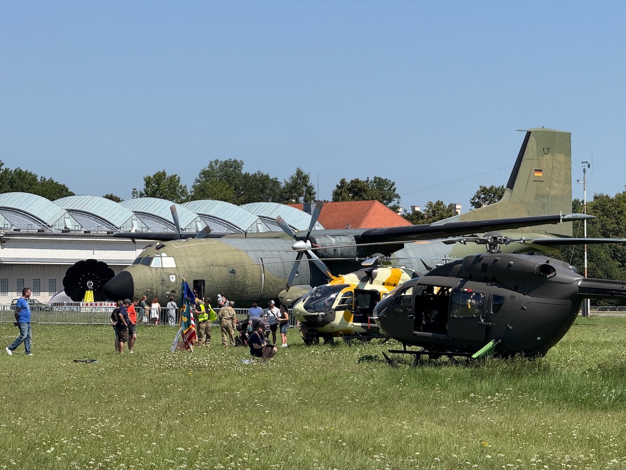 Luftfahrtmuseum Oberschleißheim – Hubschrauber und Transportflugzeug
Das Bild zeigt eine beeindruckende Szene am Flugplatz Oberschleißheim: Im Vordergrund stehen zwei Hubschrauber, umgeben von Besuchern, die das Luftfahrtmuseum erkunden. Im Hintergrund prägt eine große Transall C-160 Transportmaschine der Bundeswehr in olivgrüner Lackierung das Bild. Mit ihrem charakteristischen hochmontiertem Leitwerk und den zwei Propellern ist die Transall ein Klassiker der europäischen Luftfahrt. Die beiden Hubschrauber repräsentieren unterschiedliche Einsatztypen der militärischen Luftfahrt. Die weißen Hangars dahinter und die gepflegte Grünfläche bilden die typische Museumsumgebung. Das Bild dokumentiert die vielfältige Flugzeugsammlung des Deutschen Museums und zeigt gleichzeitig das öffentliche Interesse an historischer und moderner Luftfahrttechnik in München.